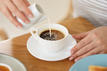 Breakfast. Woman pouring cream into cup of coffee at wooden table, closeup