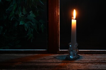 Burning candle on wooden table near window at night