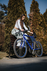 Obraz premium Outdoor portrait of a young woman seated on a bench with her bicycle, surrounded by greenery in a serene park environment.