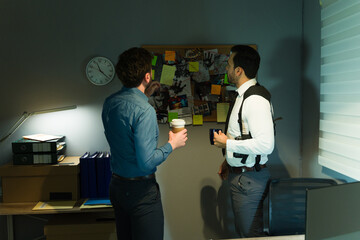 Male detectives examining a cluttered evidence board in a dimly lit room, discussing a case