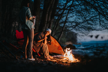 Two friends relax by a warm campfire near a tent at the shore of a lake, sharing a peaceful moment...