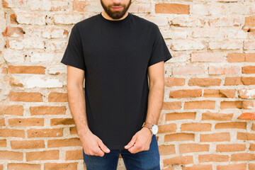 Close-up of confident man in a plain black t-shirt poses for branding mockup in front of a weathered brick wall