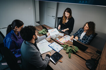A diverse group of colleagues engaged in a business meeting, collaborating and discussing project strategies using digital and physical tools in a modern office environment.