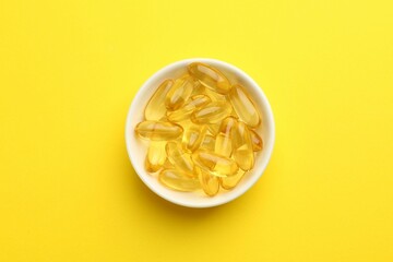 Vitamin capsules in bowl on yellow background, top view