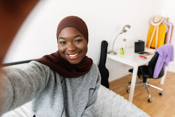 Happy young african 20s black woman in muslim headscarf taking self portrait with phone in her room