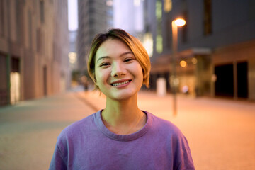 Portrait beautiful Asian nice young woman smiling with friendly expression looking joyful at camera outdoor. Generation z pretty Chinese female with blonde hair posing cheerful in city center at dusk 
