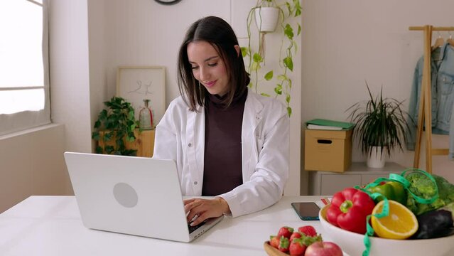 Female nutritionist in white coat working on laptop at office desk. Professional dietitian woman typing on keyboard looking at computer screen at workplace. Healthcare people concept.