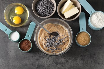 Raw dough with chocolate chips in bowl and ingredients on grey table, flat lay