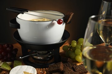 Forks with pieces of grape, bread, fondue pot with melted cheese, wine and snacks on table, closeup