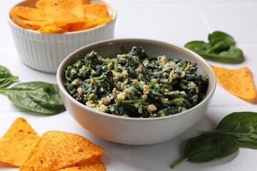 Tasty spinach dip with egg in bowl and nachos chips on white table, closeup