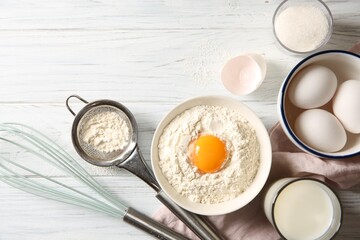 Flour with yolk in bowl and other ingredients for dough on white wooden table, flat lay