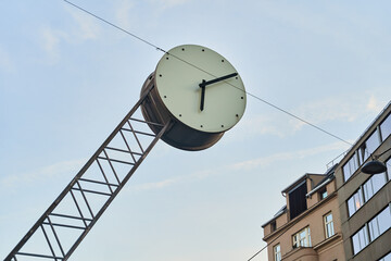 A clock hangs from a metal pole against the sky, in front of a building. The circular clock face...
