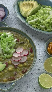 Vegan Pozole Verde dish topped with avocado, radish, and shredded napa cabbage. More toppings on the table. Vertical video.