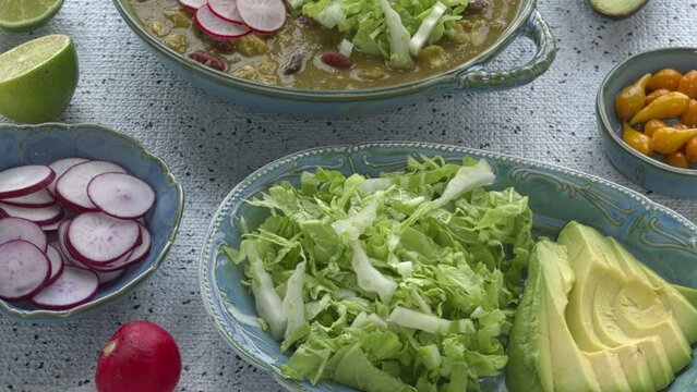 Vegan Pozole Verde dish with hominy, beans, tomatillo, and button mushrooms topped with avocado, radish, and shredded napa cabbage. More toppings on the table. Zoom in while spinning.