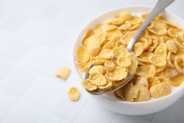 Breakfast cereal. Tasty corn flakes with milk in bowl and spoon on white tiled table, closeup. Space for text