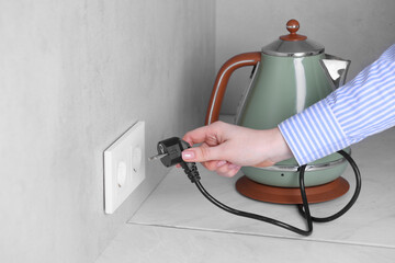 Woman plugging electric kettle into power socket at white table indoors, closeup