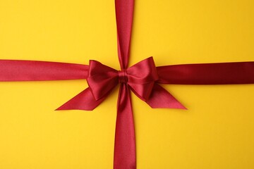 Red satin ribbon with bow on yellow background, top view