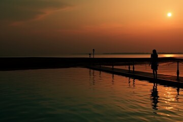 Sunset over calm lake with person standing on dock, gazing at colorful sky. Warm glow creates peaceful scene, silhouette adds mystery. Reflections of clouds on water, serene moment in nature