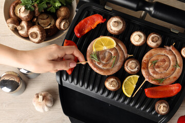 Woman cooking homemade sausages with mushrooms and bell pepper on electric grill at wooden table, top view