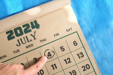 Hand pointing on July 4 2024 date on calendar flat lay in blue background. USA or United States of...