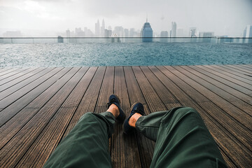 Male legs in sneakers in first person, a man sits on the roof of a skyscraper by the pool...
