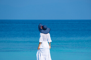 The woman wears a white Hawaiian dress and she wears a blue Hawaiian hat. She stands looking at the...