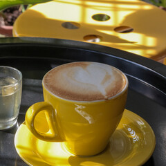A cup of coffee latte in yellow served on the round table with simple syrup. Yellow round chair and black round table. Morning Coffee. Latte art. A cup of vanilla latte served with a heart latte art.