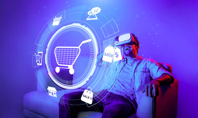 Happy man using VR headset and shopping online while sitting at sofa with shopping bags. Smart person looking at online market hologram and icon with neon background. Innovation technology. Deviation.