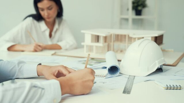 Professional architect engineer drawing blueprint and working together with young beautiful caucasian coworker on meeting table with safety helmet, house model, blueprint scatter around. Immaculate.