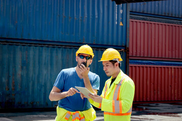 Asian engineer worker man with safety helmet and protective clothing holds tablet and discuss with his colleagues who holds walkie talkie. Male worker working at logistic shipping container cargo yard