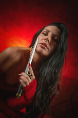 Obraz premium Young beautiful girl with dark hair and in the red dress posing in the dark background with the dagger sword in hands.