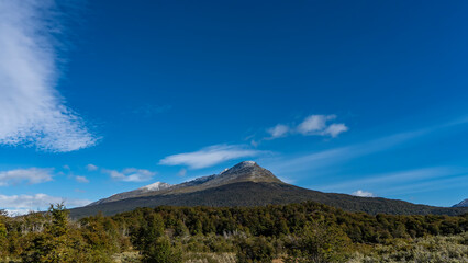 The beautiful landscape of Patagonia. Snow-capped mountain on a background of blue sky and clouds. In the foreground is a valley overgrown with green forest. Argentina. Tierra del Fuego National Park.