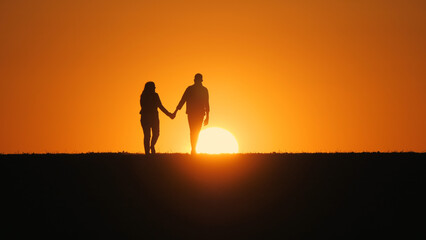 A young couple walks across a field towards the setting sun, holding hands