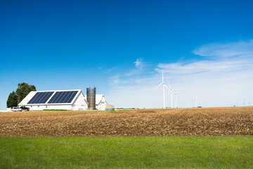 A vibrant landscape featuring a farm with a large solar panel array and several wind turbines against a clear blue sky, set in a plowed field.