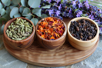 Three bowls of spices are arranged on a wooden tray. The bowls contain cinnamon, cloves, and nutmeg. The spices are arranged in a way that they are not overlapping each other