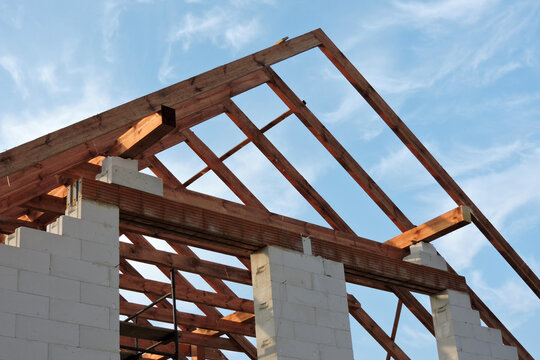 A timber roof truss in a house under construction, walls made of autoclaved aerated concrete blocks, rough window openings, a reinforced brick lintel, a scaffolding, blue sky in the background