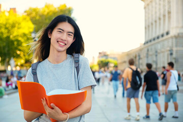 Taiwanese male student smiling and looking at camera with a folder and backpack at university campus in Spain.