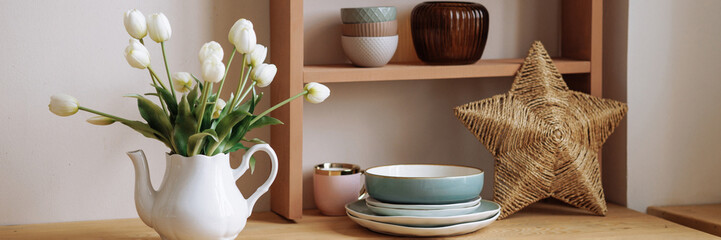 Stylish interior of a wooden Scandinavian kitchen. Fresh flowers in a vase. Ceramic plates, dishes, utensils and cozy decor on wooden shelfs. Open shelves in the kitchen. Banner with copy space.
