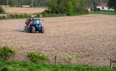 Blue farm tractor pulls disc harrow cultivator over agricultural field with rolling hills background