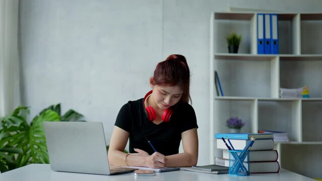 Young woman student listen music in headphone and writing on note book about lists works things to do homework