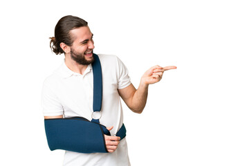 Young handsome man with broken arm and wearing a sling over isolated chroma key background pointing...
