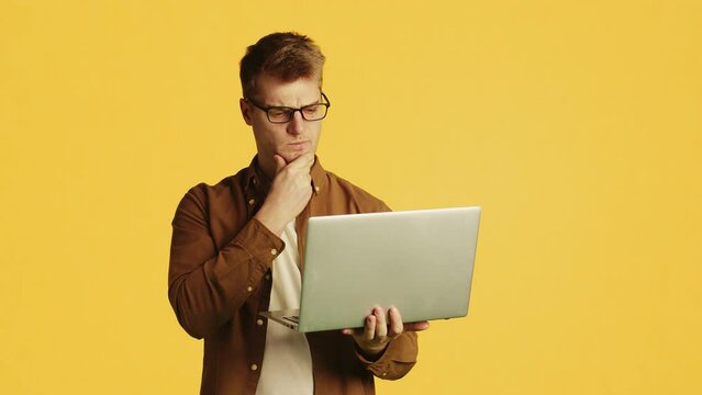 Young attractive caucasian man in glasses and casual clothes standing with a laptop, thinking with a perplexed face, stroking his chin with a hand on a yellow background in a studio
