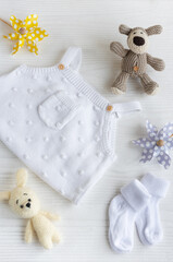 Set of white clothes and accessories for newborn baby. Knitted toys, knitted romper, socks on white wooden background.