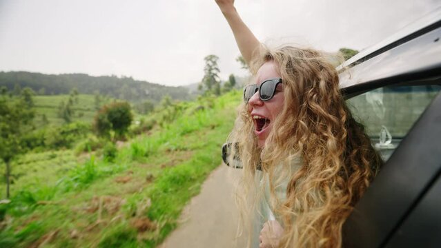 Cheerful woman with curly hair enjoys mountain road trip in car. Smiling female traveler leans out window, feels wind, adventure in nature. Joyful road trip, freedom in wild landscape, hair blowing.