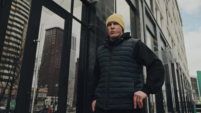 Person with a focused expression wearing a yellow beanie and black puffer vest, walking in front of a glass-walled urban building.