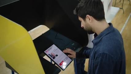 Caucasian man makes choice and votes for US presidential candidate in voting booth using tablet computer. US citizen at polling station during National Election Day in the United States. High angle.