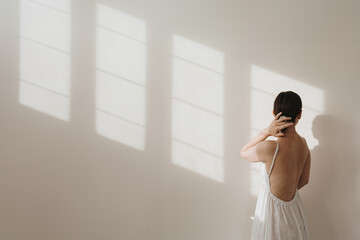 Young pretty woman in linen dress over white wall with soft sunlight shadows. Silhouette in sunlight. Aesthetic fashion vogue concept