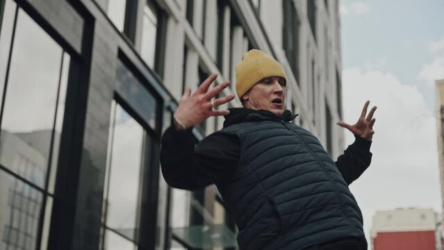 Person wearing a yellow beanie and black puffer vest walking energetically in an urban setting with modern glass buildings.