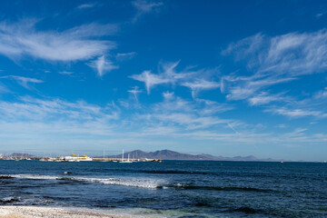 Fototapeta premium Walking on sea promenade in Corralejo along black rocks, blue water, view on Lanzarote island, Fuerteventura, Canary islands, Spain