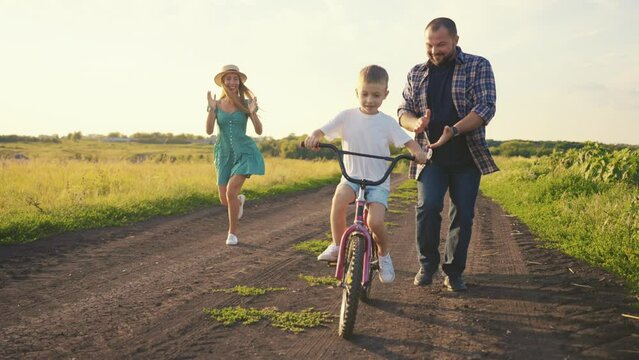 Mom dad teaching son ride bike on rural road in village on vacation. Parents running near child boy, mother holding bike steering wheel, father supporting kid. Family happy weekend on nature.